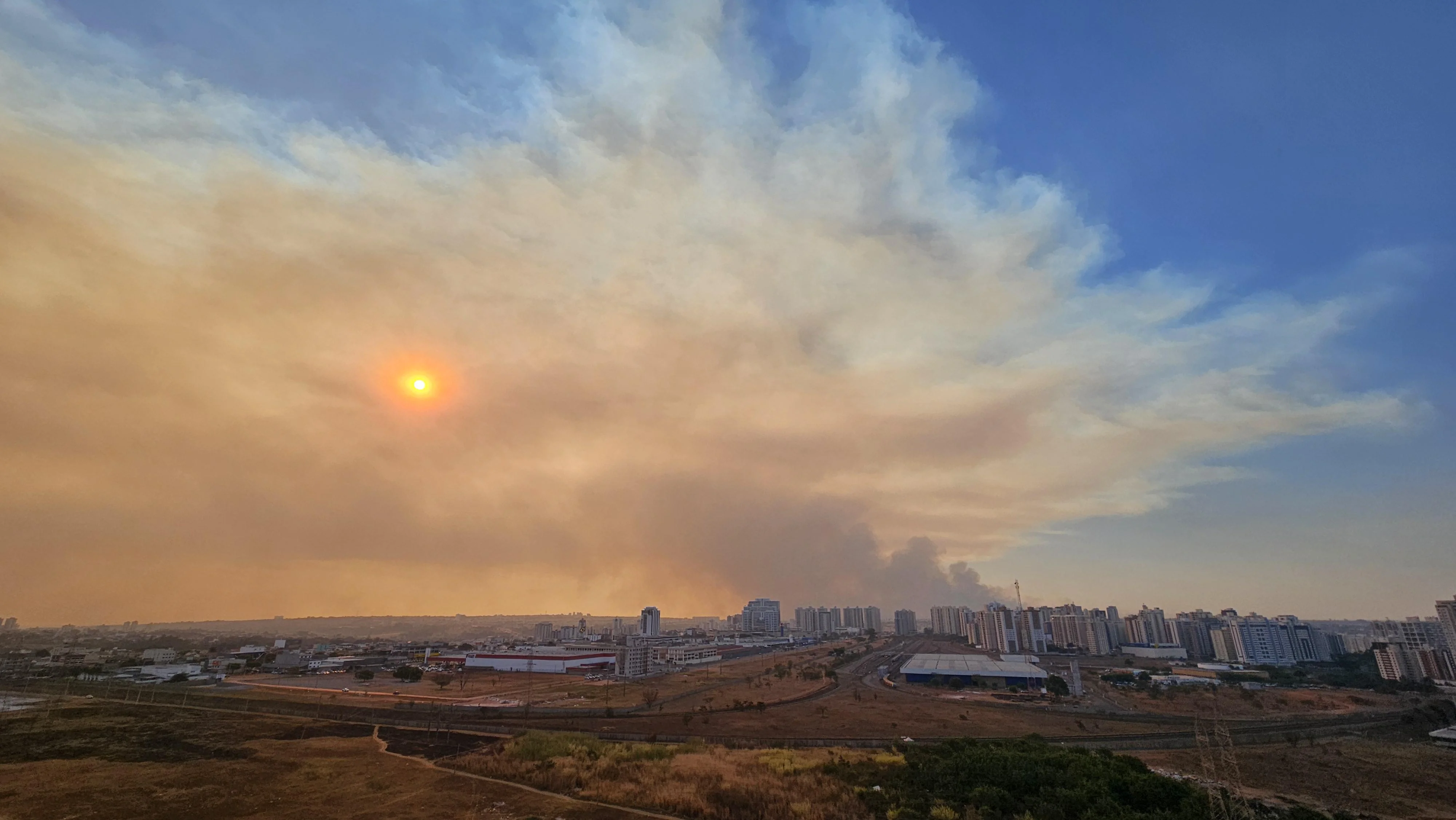 Vista do horizonte de Águas Claras: o céu está bem azul, mas um foco de fumaça está se espalhando, cobrindo o céu e o sol, que apresenta um aspecto alaranjado.