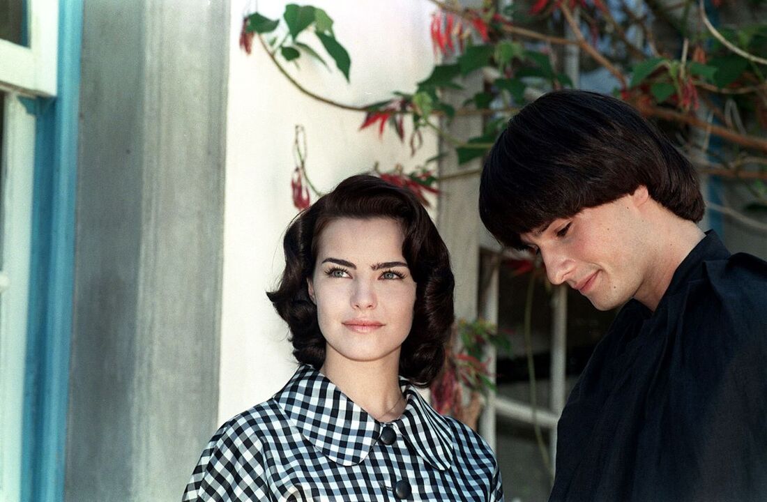   Hilda Furacão, a woman in a checkered outfit stands outdoors next to a man, Malthus, in a dark shirt, who is looking down. 