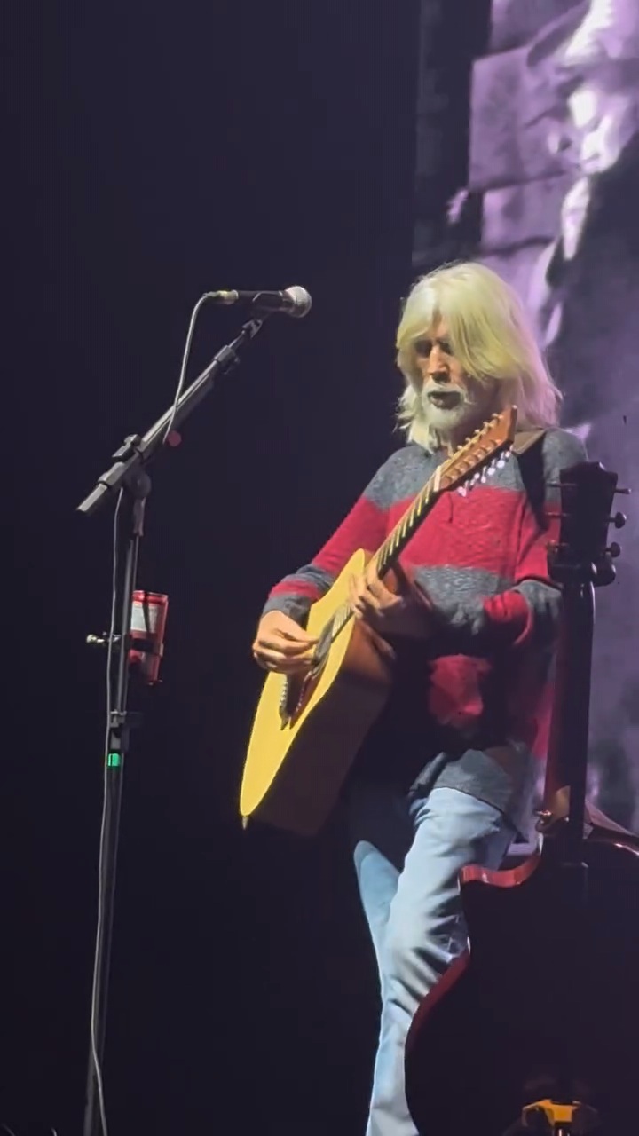 Oswaldo Montenegro plays an acoustic guitar on stage, wearing a red and gray sweater and jeans, with a microphone nearby.